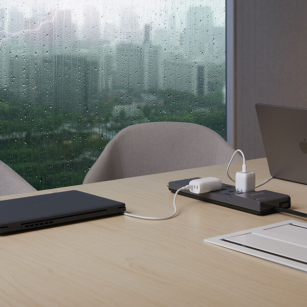 A modern workspace with a wooden table featuring a sleek power strip charging multiple devices, including a laptop and an adapter. A large window in the background shows a rainy cityscape with raindrops on the glass, creating a calm and productive atmosphere.