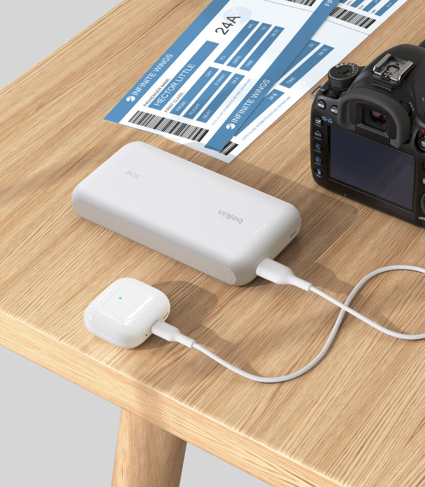 A white Belkin BoostCharge Power Bank 20K with Integrated Cable is placed on a wooden table, charging a pair of white AirPods, with a DSLR camera and boarding passes nearby. A white Belkin BoostCharge Power Bank 20K with Integrated Cable is placed on a wooden table, charging a pair of white AirPods, with a DSLR camera and boarding passes nearby.