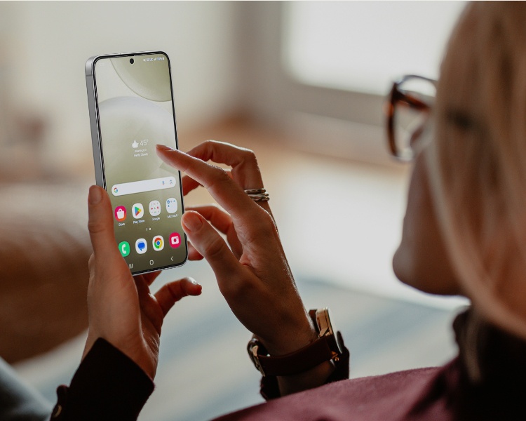 Cette image montre une personne assise sur un canapé en train d'utiliser son smartphone. L'attention est portée sur l'écran du téléphone, qui est clair et net, ce qui suggère l'utilisation d'une protection d'écran. La personne semble détendue, ce qui met en avant le côté confortable et utile de la protection d'écran. L'arrière-plan est floue avec une lumière naturelle chaude qui créé une ambiance chaleureuse et met en avant l'appareil avec son écran clair et net.