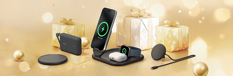 Holiday-themed banner featuring Belkin tech products on a gold background, including pink over-ear headphones, a wireless charging stand holding a phone, smartwatch, and earbuds, and a gray Belkin carrying case, with a wrapped gift in the background.