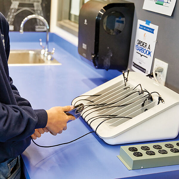 A student unplugs a smartphone from a multi-device charging station on a blue countertop. The station has several phones charging in individual slots, and a sign on the wall nearby promotes ordering a yearbook.