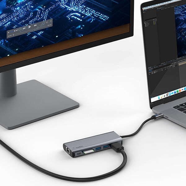 A modern workstation featuring a MacBook connected to a Belkin USB-C hub, which links the laptop to an external monitor displaying a circuit board graphic. The hub also provides additional connectivity options, including Ethernet and USB ports, with neatly arranged cables on a clean white desk.
