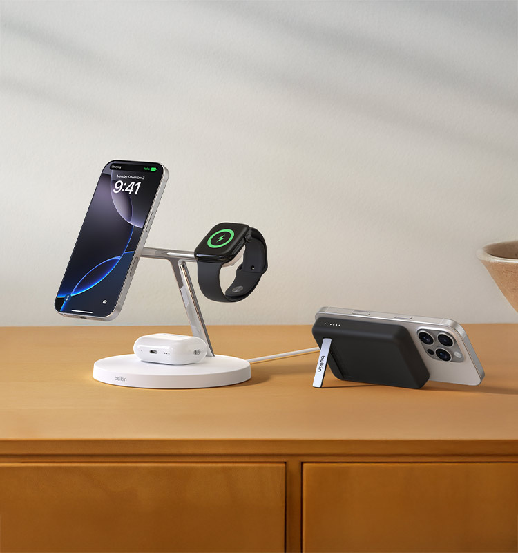 A white Belkin BoostCharge Pro 3-in-1 Wireless Charger with Official MagSafe Charging 15W and a black Belkin BoostCharge Magnetic Wireless Power Bank 5K + Stand are displayed on a wooden surface, charging an iPhone, Apple Watch, and AirPods.