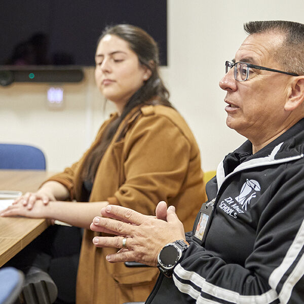 Two individuals seated at a conference table in discussion; the man in the foreground wears glasses and a Da Vinci Schools jacket, gesturing as he speaks, while the woman in the background listens attentively.