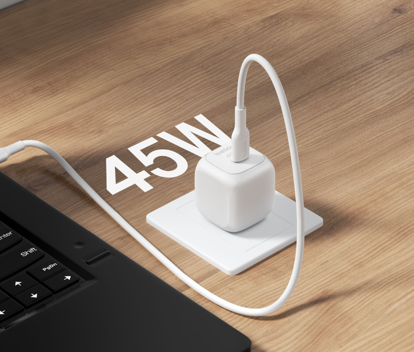 A Belkin BoostCharge Compact USB-C Wall Charger 45W is plugged into a wall socket on a wooden surface, charging a connected laptop, with "45W" displayed in bold text.