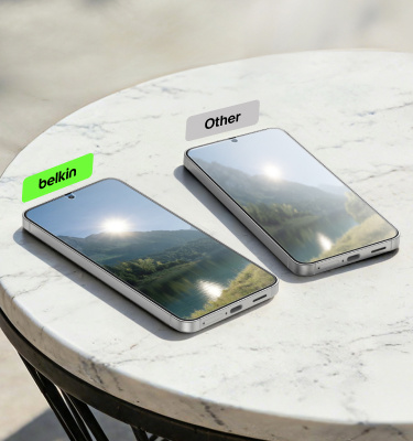 Two smartphones resting on a round marble table in bright sunlight, one labeled &lsquo;belkin&rsquo; and the other &lsquo;Other,&rsquo; comparing reduced glare versus stronger screen reflections.