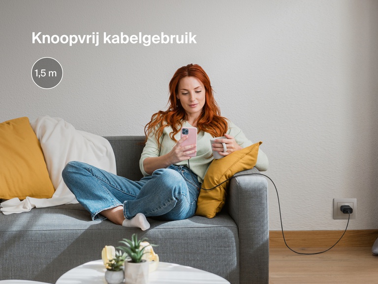 Een vrouw zit comfortabel op de bank terwijl ze haar iPhone gebruikt, die is aangesloten via een USB-A-kabel met Lightning-connector. De kabel van 1,8 meter blijft knoopvrij en is extra lang voor flexibiliteit bij het opladen.