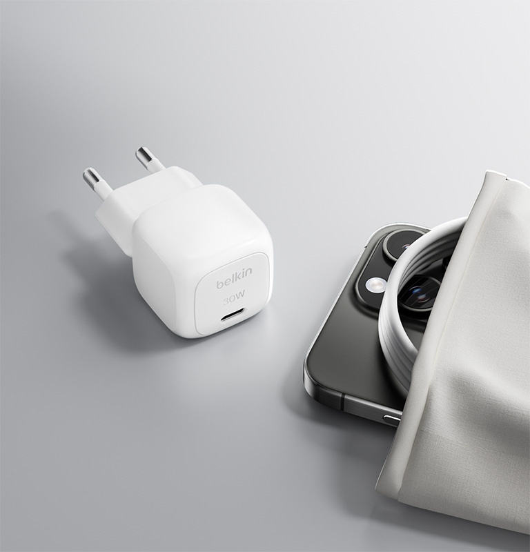 L'image montre le chargeur mural USB-C BoostCharge Compact 30 W de Belkin avec des broches pliables placées dans une valise à côté d'un passeport et d'un appareil photo, soulignant son design compact et adapté aux voyages.