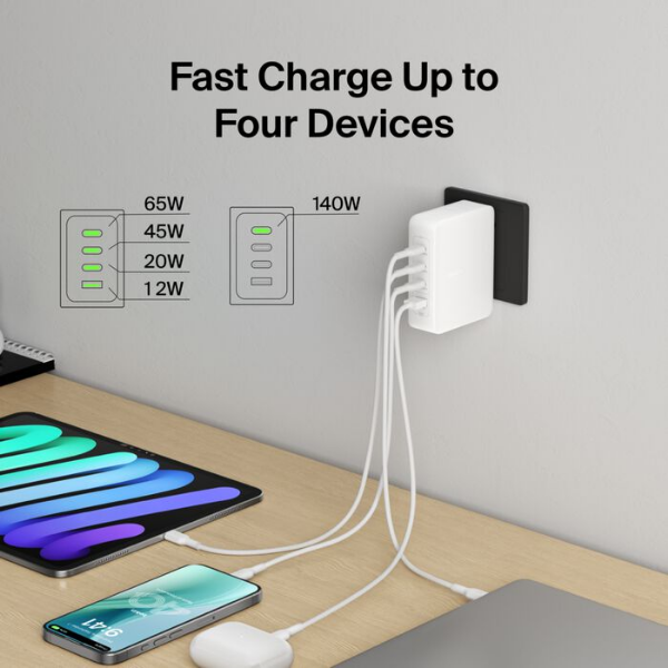 A wall-mounted white multi-port charger plugged into an outlet, charging a laptop, tablet, smartphone, and earbuds simultaneously. On-screen text reads &lsquo;Fast Charge Up to Four Devices&rsquo; with power output icons, and the devices rest on a desk below connected by white cables.