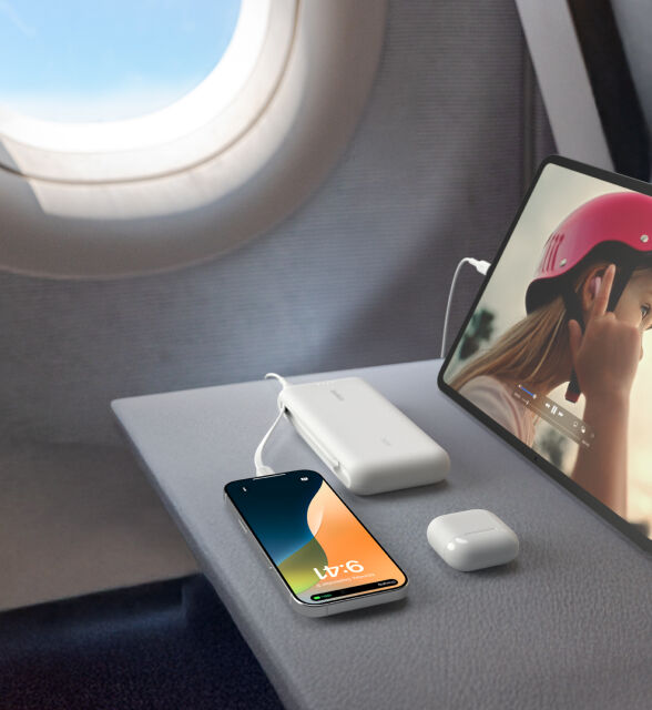 A white Belkin BoostCharge Power Bank 20K with Integrated Cable is placed on an airplane tray table, charging an iPhone and iPad, with a pair of AirPods nearby. A white Belkin BoostCharge Power Bank 20K with Integrated Cable is placed on an airplane tray table, charging an iPhone and iPad, with a pair of AirPods nearby.