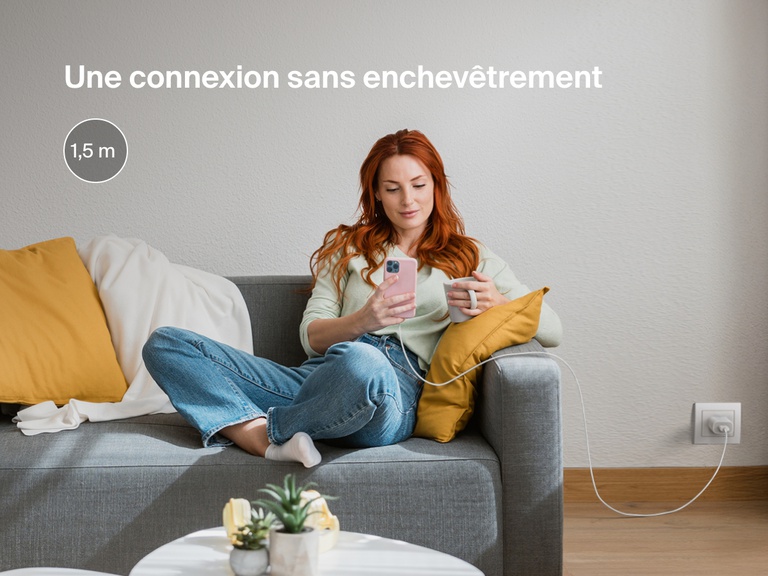 Une femme confortablement assise sur un canapé utilise son iPhone connecté à un câble USB-A Belkin BoostCharge avec connecteur Lightning de 1,8 m de long pour une recharge sans nœud, même loin d'une prise.