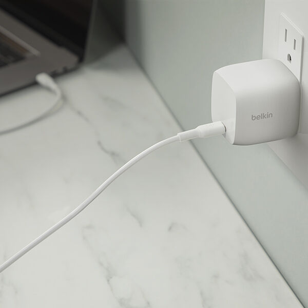 Close-up of a white Belkin wall charger plugged into an outlet, with a charging cable extending across a light marble desk surface toward a laptop in the background.