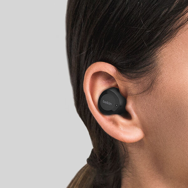 Close-up of a person's ear wearing a black Belkin wireless earbud. The earbud has a sleek, compact design with the Belkin logo visible on the outer surface, fitting snugly in the ear. The person has dark hair, and the background is a neutral gray.