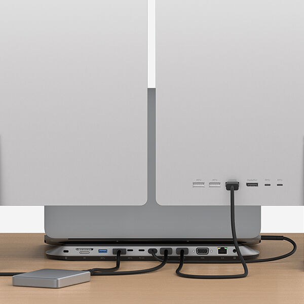 A dual-monitor workstation setup viewed from the back, featuring a slim docking station with multiple connected cables. The docking station facilitates various connections, including HDMI, VGA, USB, and Ethernet. A compact external hard drive is also connected, demonstrating an efficient cable management system for a professional workspace.