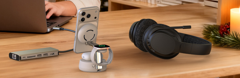 Holiday banner featuring Belkin home office tech gifts on a wooden desk, including a USB-C hub, a wireless charging stand holding a phone and smartwatch, and a black headset, with a lit candle and festive greenery in the background.