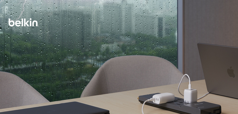 A modern workspace with a wooden table near a large window covered in raindrops, showing a blurred cityscape in the background. The table has a Belkin power strip with multiple devices plugged in, including a white charger and a MacBook laptop. Another closed laptop is also on the table. The setting has a calm and professional atmosphere, with a focus on productivity and connectivity. The "Belkin" logo is visible in the top left corner.