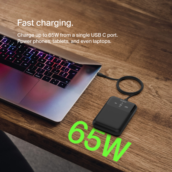 This image shows a laptop on a wooden surface, connected to a compact power bank via a USB-C cable. The text overlay highlights the product's capability for "Fast charging," stating it can charge up to 65W from a single USB-C port, suitable for powering phones, tablets, and even laptops. The number "65W" is prominently displayed in bright green, emphasizing the charging power. The laptop's keyboard features colorful lighting, adding a modern and tech-savvy aesthetic.