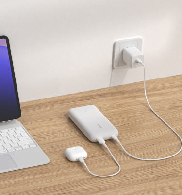 白いBelkin BoostChargeモバイルバッテリー20K（一体型ケーブル付き）が木製の表面に置かれ、壁のコンセントから充電しながらAirPodsを1組充電しており、近くにはタブレットがあります。