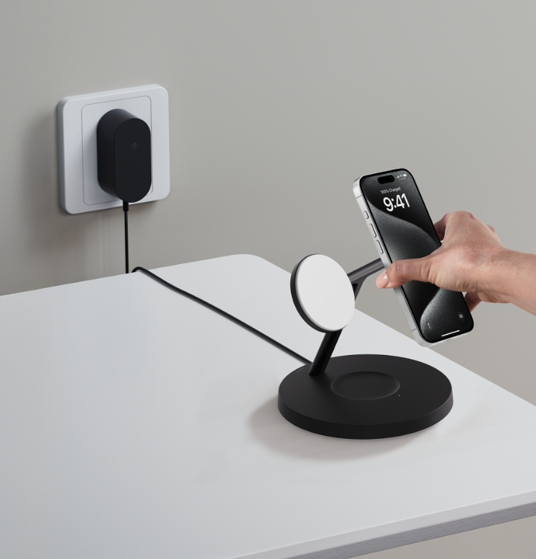 A person sets an iPhone on a black BoostCharge Pro 3-in-1 Wireless Charging Stand with Qi2 15W, the included 40W PSU is visible in the background plugged into a power outlet. 