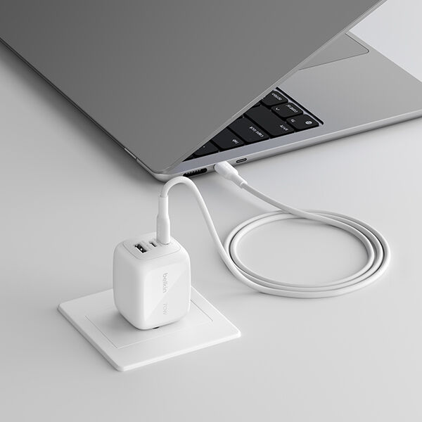 A white wall charger plugged into a desk outlet, connected by a cable to a partially open laptop resting on a clean, light-colored surface.
