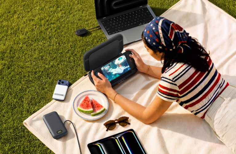 Woman relaxing outdoors on a picnic blanket while gaming on a handheld device surrounded by Belkin tech accessories and a laptop.