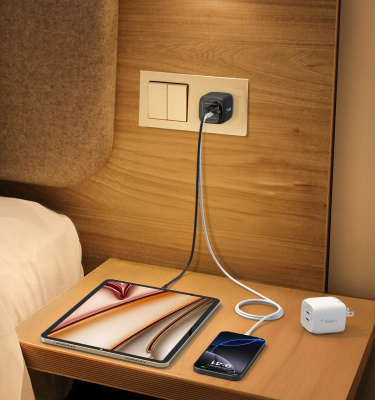 An iPad and iPhone charge on a bedside table using the Belkin Secure AirTag Holder Bundle with dual-port charger and braided cables, ideal for organized travel and efficient charging.