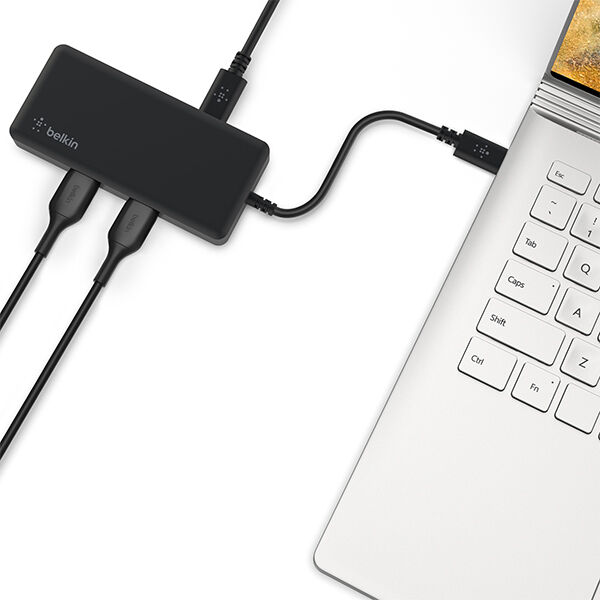 Close-up of a black Belkin docking hub connected to a silver laptop via USB-C, with multiple cables plugged into the hub on a white background.