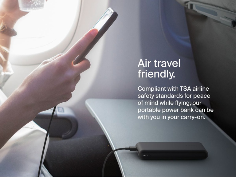 A person uses a black Belkin BoostCharge Power Bank 20K to charge their phone on an airplane tray table, highlighting its TSA-compliant, travel-friendly design.