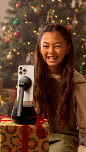 A young girl smiles while video chatting on an iPhone mounted on a Belkin Stage Auto-Tracking Stand Pro with DockKit, placed on a festively wrapped gift box in front of a decorated Christmas tree.