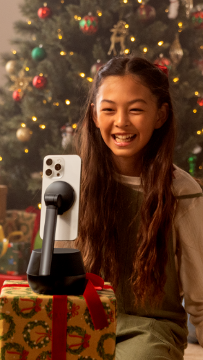 Una giovane ragazza sorride durante una videochiamata tramite un iPhone montato sul Supporto Pro con tracking automatico compatibile con DockKit Stage di Belkin; il supporto posizionato su un pacco regalo con carta festiva, di fronte a un albero di Natale decorato.