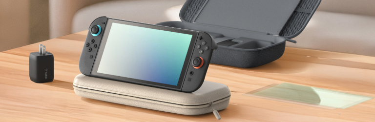 A Nintendo Switch 2 console is propped on a light-colored protective carrying case atop a wooden desk. Nearby, a black wall charger stands upright, and an open dark gray storage case with compartments is visible in the background. A screen protector lies flat on the desk, completing the setup of essential gaming accessories in a clean, well-lit room.