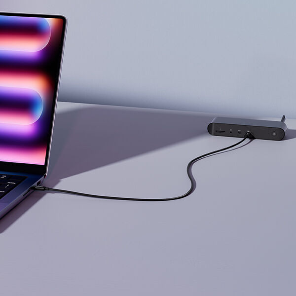 A laptop connected to a compact Belkin docking station via a single black cable, placed on a minimalist purple desk with a clean background.
