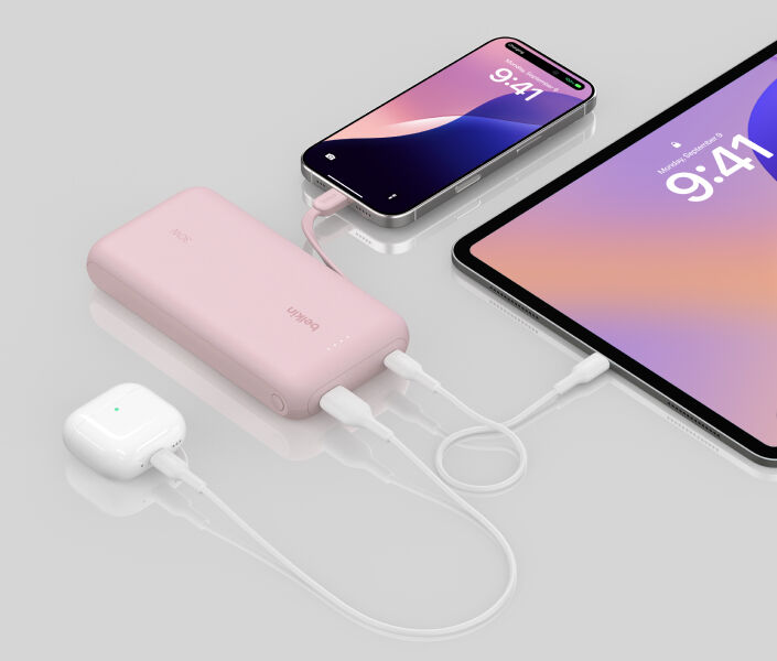 A pink Belkin BoostCharge Power Bank 20K with Integrated Cable is shown charging an iPhone, an iPad, and AirPods simultaneously on a reflective surface.