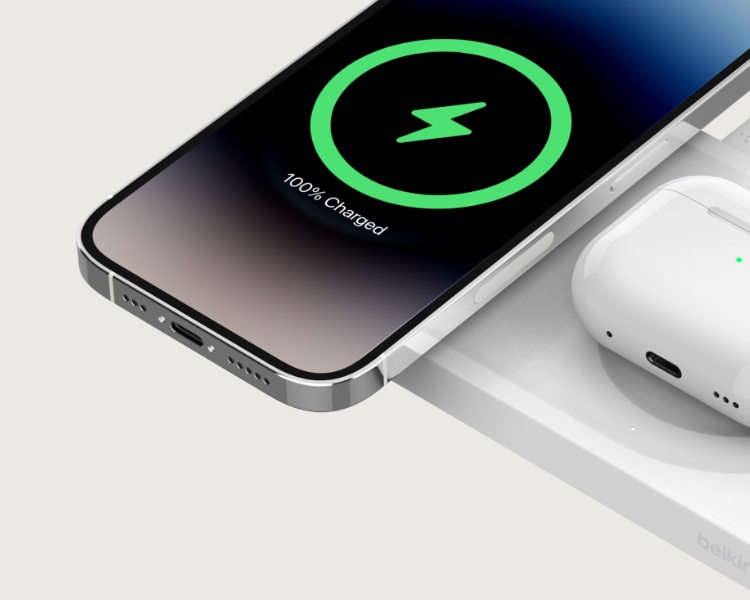 Ein Smartphone und ein kabelloser Kopfhörer, die auf einem kabellosen MagSafe-kompatiblen Ladegerät von Belkin aufgeladen werden. Auf dem Handy-Bildschirm ist ein grünes Ladesymbol mit dem Text „100% Charged“ zu sehen, wodurch angezeigt wird, dass der Akku vollständig aufgeladen ist.
