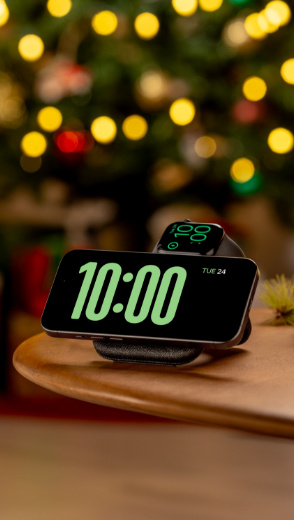 An iPhone in StandBy mode and an Apple Watch are displayed charging on a Belkin 2-in-1 Magnetic Charging Travel Pad with Qi2 15W, set against a festive backdrop of a Christmas tree with glowing lights, creating a warm holiday ambiance.