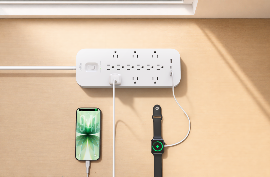 Belkin surge protector with AC outlets and USB-C ports charging a smartphone and smartwatch on a tabletop, providing convenient power and surge protection for everyday devices.