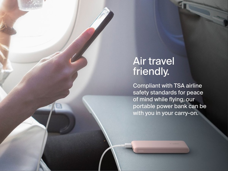 A person uses a smartphone on an airplane while a Belkin BoostCharge Power Bank 10K rests on the tray table, illustrating its TSA-compliant, travel-friendly design for carry-on use.