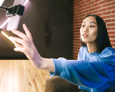 A woman in a blue sweatshirt reaches out toward a smartphone mounted on a stand with a glowing LED light setup in the background. She is seated at a wooden table with a brick wall and dark door behind her.