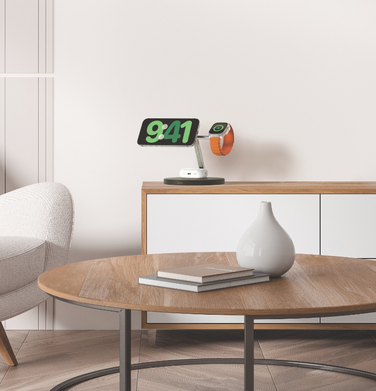 An iPhone in StandBy mode, AirPods, and an Apple Watch are shown charging on a Belkin BoostCharge Pro 3-in-1 Wireless Charging Stand with Qi2 15W, placed on a wooden cabinet in a modern, minimalist living room, with a round wooden coffee table and a decorative vase in the foreground.