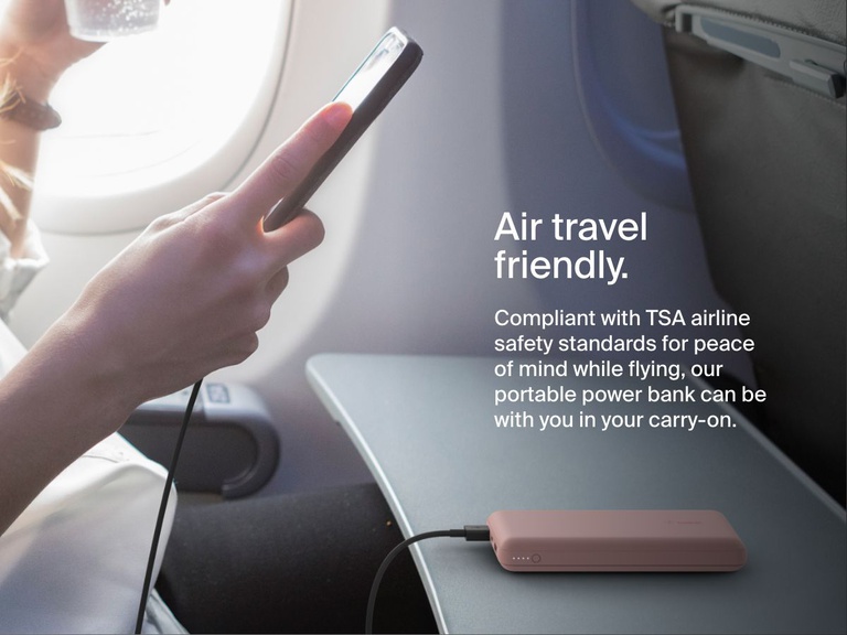 A person uses a Belkin BoostCharge Power Bank 20K to charge their phone on an airplane tray table, highlighting its TSA-compliant, travel-friendly design.