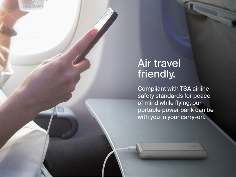 A person uses a silver Belkin BoostCharge Power Bank 20K to charge their phone on an airplane tray table, highlighting its TSA-compliant, travel-friendly design.