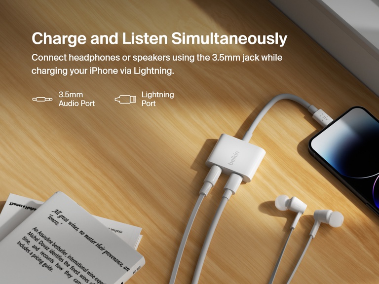 Belkin 3.5mm Audio + Charge RockStar™ connected to an iPhone enables simultaneous charging via Lightning and audio playback through the 3.5mm jack, shown on a wooden desk with earphones and reading materials.