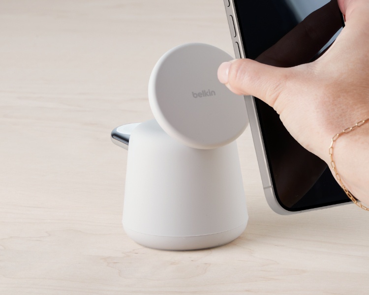 Die Hand einer Person hält ein iPhone in der Nähe einer weißen Belkin MagSafe-Ladestation auf einer hellen Holzoberfläche. Die Ladestation hat eine modernes, minimalistisches Design mit einem runden, magnetischen Ladepad, das so ausgerichtet ist, dass das Telefon leicht darauf platziert werden kann. Die Person trägt ein filigranes goldenes Armband.