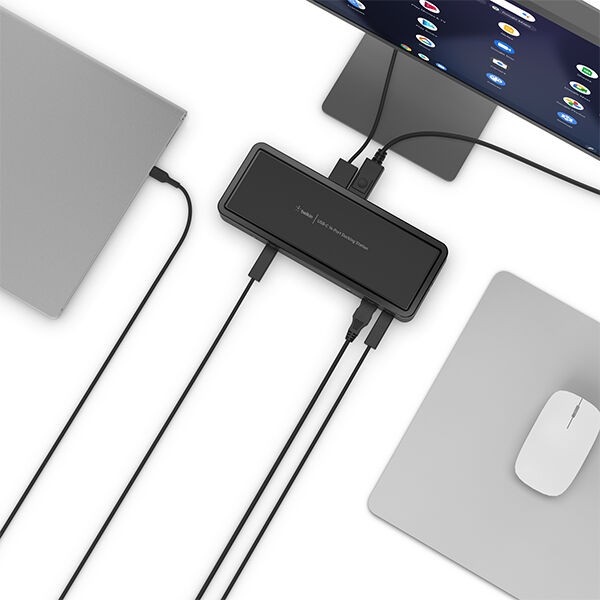 Top-down view of a clean desk setup showing a laptop connected to a compact docking hub, with multiple cables running to an external monitor and peripherals, alongside a mouse on a mouse pad.