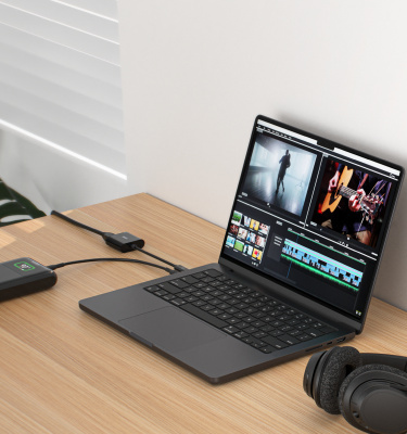 Remote work setup featuring a laptop displaying video editing software, connected to an external power bank via a USB-C adapter. Over-ear headphones rest on the wooden desk, with a plant and window blinds in the background, creating a comfortable workspace.