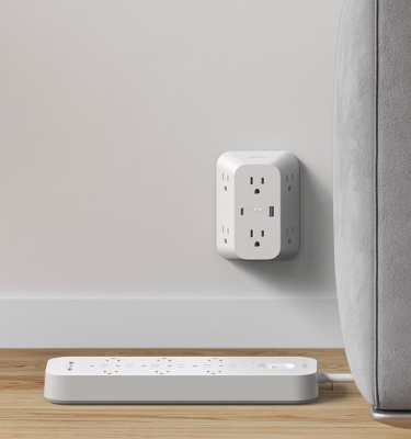 Wall-mounted Belkin Surge Protector with multiple outlets is shown in use beside a gray sofa, with a power strip resting on the wooden floor nearby.