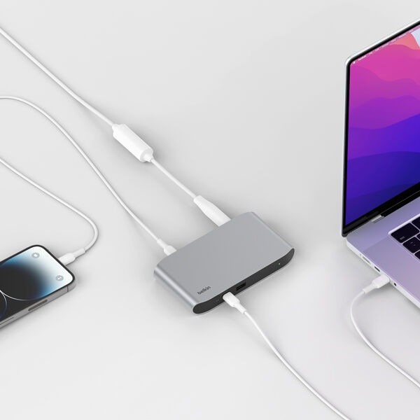 A Belkin USB-C hub connected to a MacBook and iPhone, enabling multi-device charging and data transfer. The sleek, silver hub is positioned on a clean, white surface, demonstrating its compact and modern design. Multiple cables are plugged into the hub, showcasing its versatile connectivity options, including USB-C pass-through charging and additional ports for accessories.