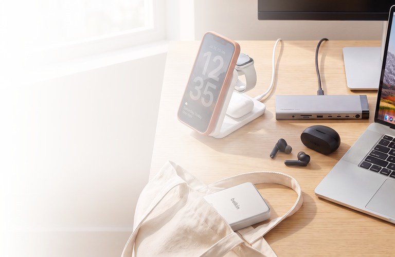 A sunlit desk with an iPhone in an orange case on a Belkin 3-in-1 magnetic charging stand beside an Apple Watch and AirPods, a Belkin Thunderbolt dock wired to a laptop, black earbuds, and a Belkin power bank in a canvas tote