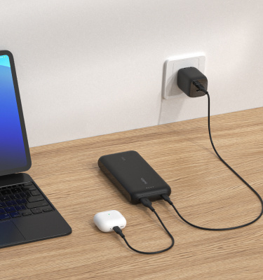 A black Belkin BoostCharge Power Bank 20K with Integrated Cable is placed on a wooden surface, charging a pair of AirPods while being recharged from a wall outlet, with a tablet nearby.