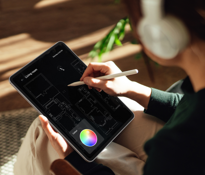 Una persona con auriculares utiliza una tableta con un lápiz Apple para diseñar o editar un plano digital. La pantalla muestra un plano detallado del piso con una herramienta de selección de color abierta en la esquina. La luz del sol ilumina parte de la escena, creando una atmósfera cálida y enfocada.